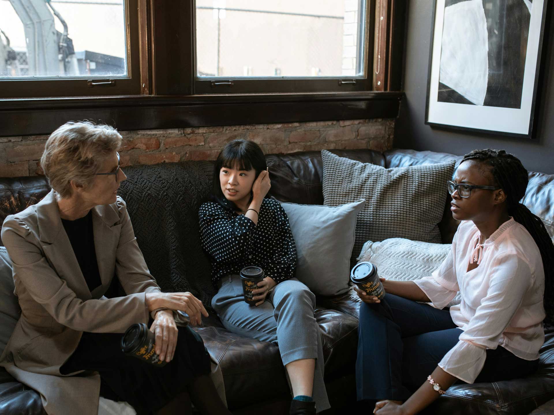 sThree women sit on a sofa holding coffee cups and having a discussion