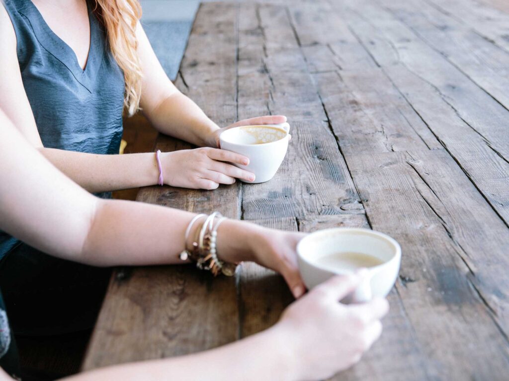 two women sitting at a table hold coffee cups while they talk