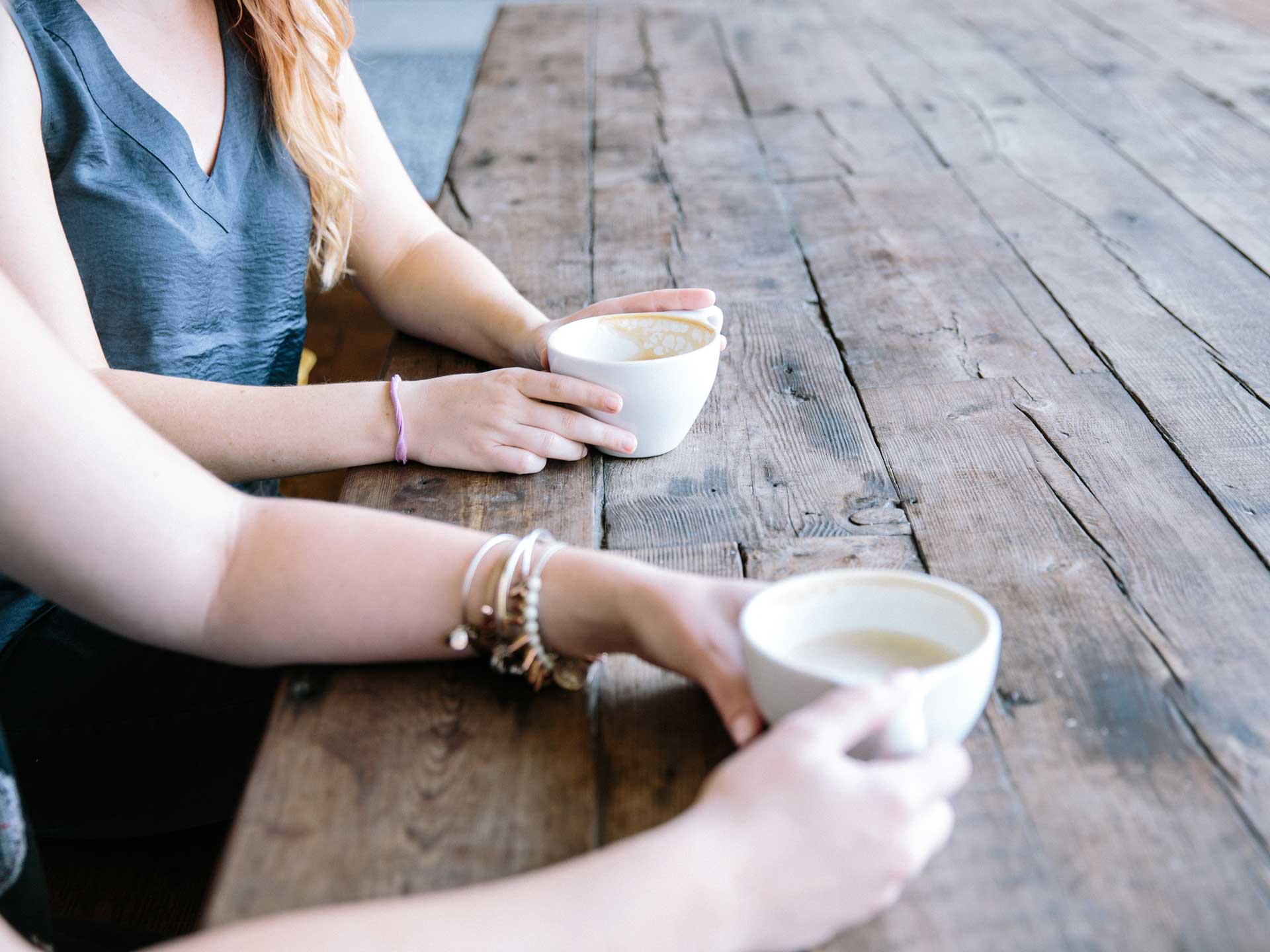 two women sitting at a table hold coffee cups while they talk