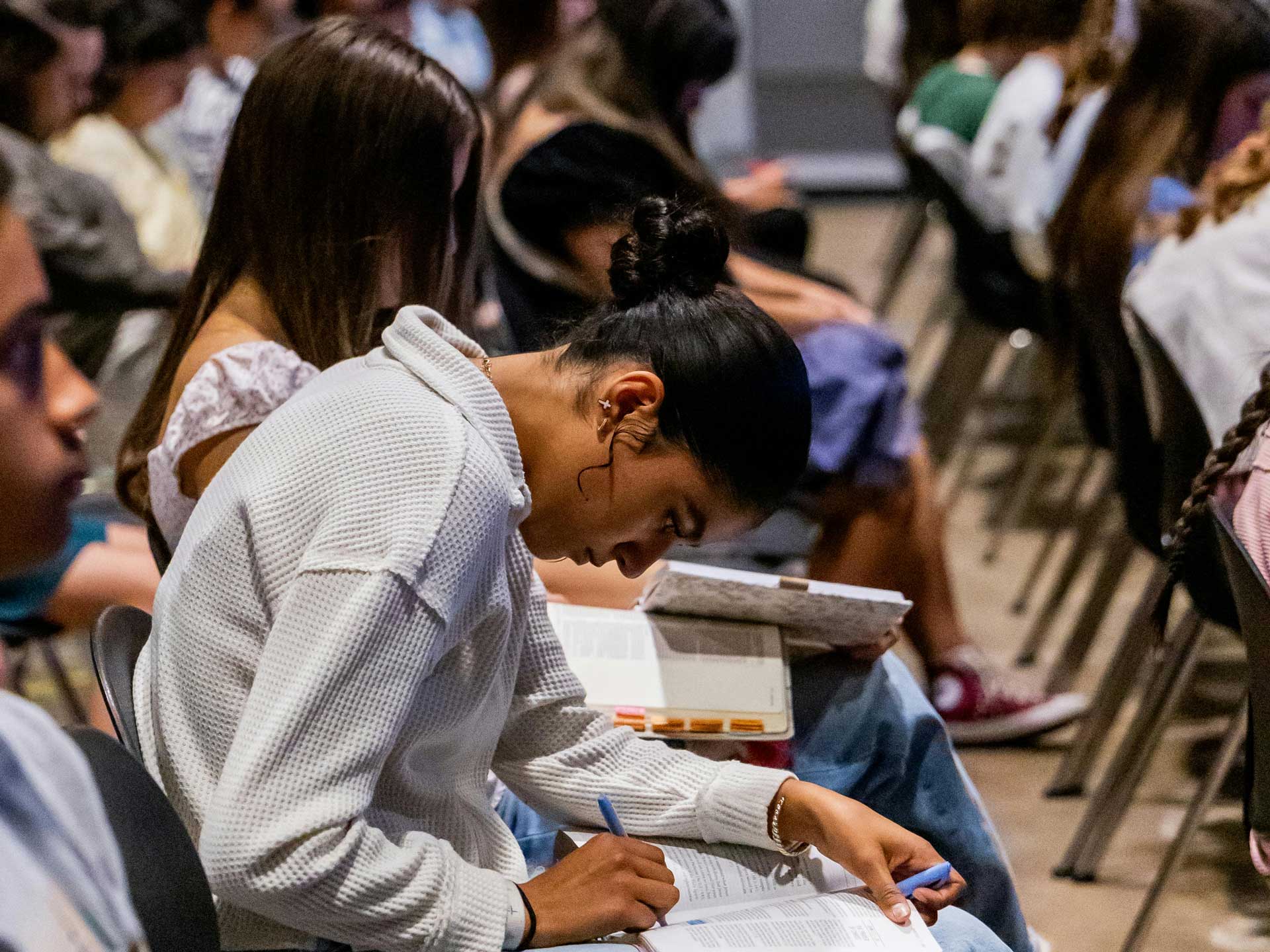 A woman sitting in the audience at a lecture bending forward to write notes in a pad on her knee