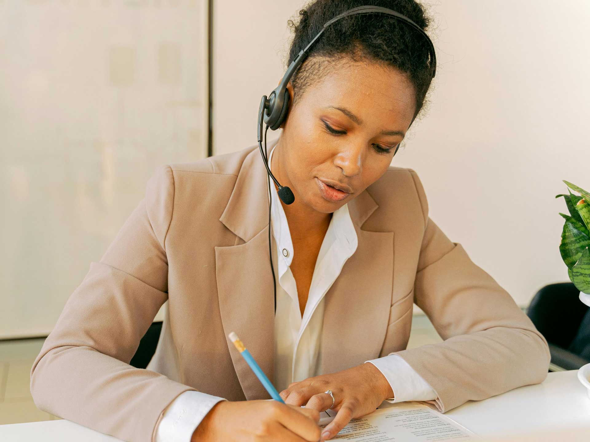 A woman in a suit sits at a desk wearing a headset. She is writing notes on a pad with a pencil.
